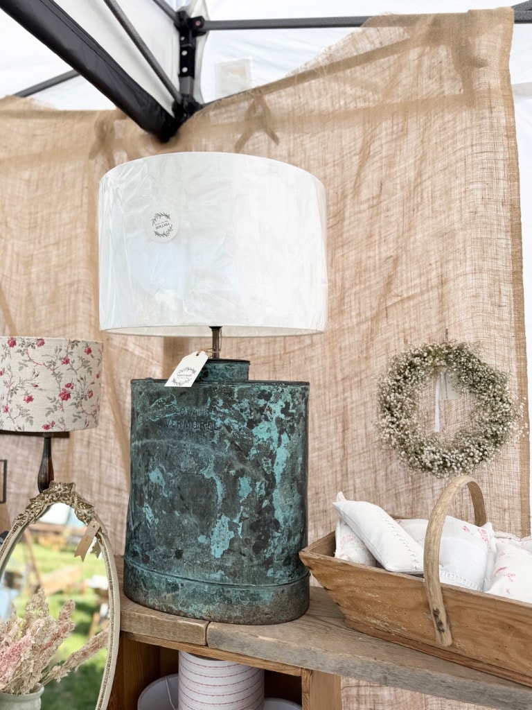 Antique verdigris copper vessel lamp with white drum shade at vintage fair stall, dried flower wreath and floral lampshade in background